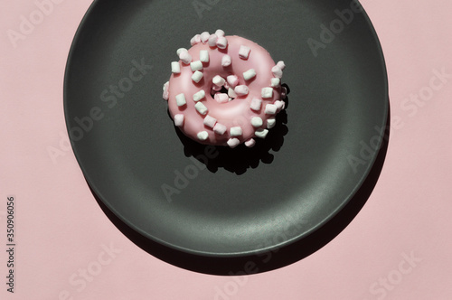 One pink donat with white marshmallows on the dark grey ceramic plate on the light coral background. Geometric sunny creative trendy composition with shadows. Art idea for celebration donat day photo