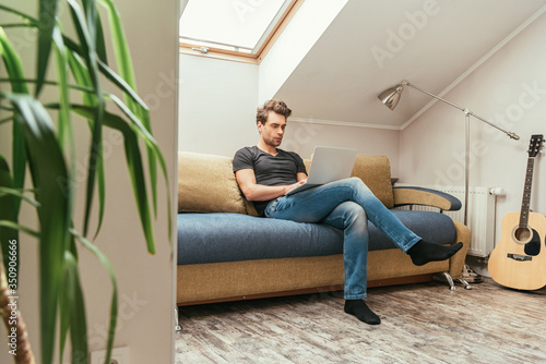 Photos selective focus of handsome man using laptop while sitting on sofa in attic room