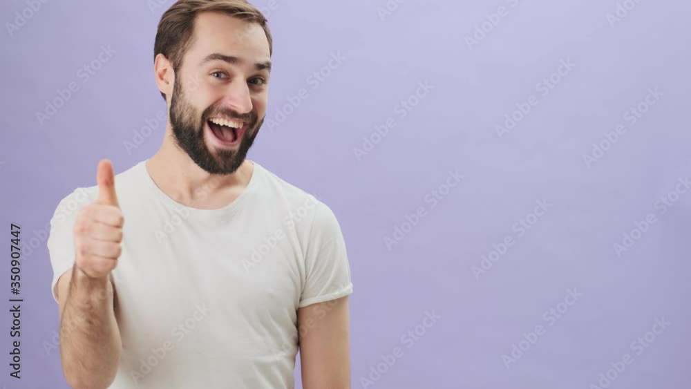 A happy young man wearing a white t-shirt is pointing to the side and showing thumb up gesture standing isolated over gray background A happy young man wearing a white t-shirt is pointing to the side and showing thumb up gesture standing isolated over gray background