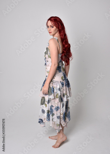 Portrait of a beautiful woman with red hair wearing a flowing floral gown. full length standing pose, isolated against a studio background