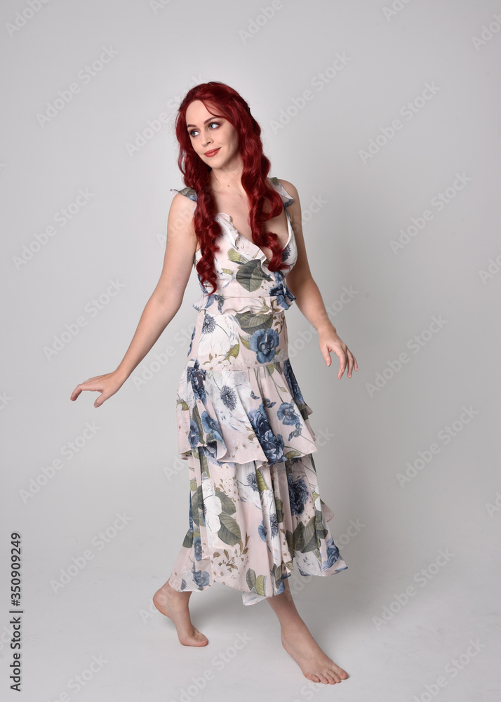 Portrait of a beautiful woman with red hair wearing a flowing floral gown. full length standing pose, isolated against a studio background Portrait of a beautiful woman with red hair wearing a flowing floral gown. full length standing pose, isolated against a studio background