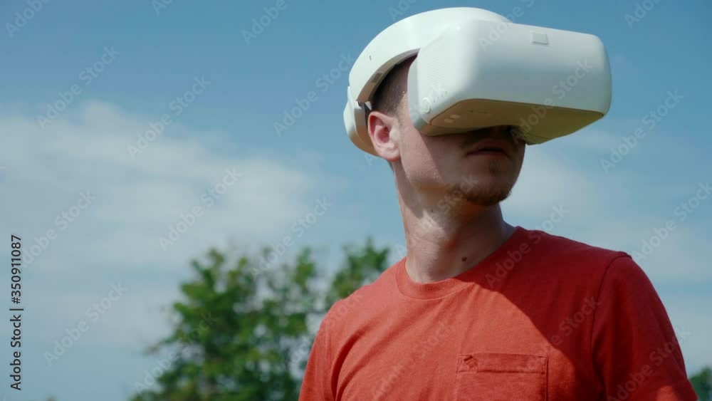 Man in a virtual reality helmet is studying virtual game. The guy in the park in a red T-shirt on a background of blue sky and green trees looks VR glasses. 4K footage Man in a virtual reality helmet is studying virtual game. The guy in the park in a red T-shirt on a background of blue sky and green trees looks VR glasses. 4K footage