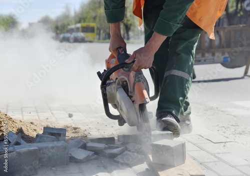 A worker saws a small sidewalk block with a saw,stepping on it with his foot.