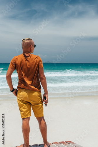 A young guy in a brown T-shirt and shorts stands on the sandy seashore in sunglasses. Clear summer day, blue sky with white clouds, waves with white foam