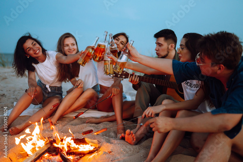Group of friends near a campfire on the beach cheers and drinking beer at night. Summer holidays, vacation, relax and lifestyle concept. © maxbelchenko