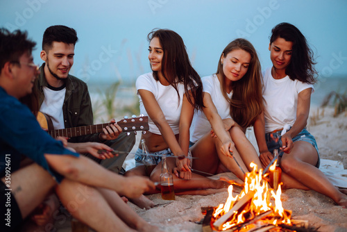 Group of young friends sitting on beach and fry sausages. One man is playing guitar. Summer holidays, vacation, relax and lifestyle consept. Camping time. © maxbelchenko