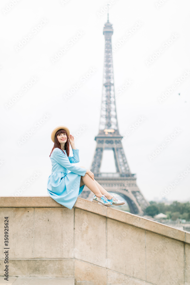 Beautiful parisian red haired girl in stylish hat and blue dress sitting at the stairs with the Eiffel tower background, and looking up with nice smile