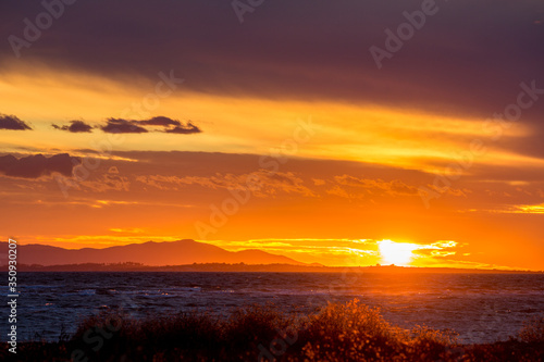 Sun is setting down to the West partially hidden behind curly clouds near the fishermen village of Porto Lagos  Northern Greece. Mountain range and sea water in darkness. Blurred bushes in foreground