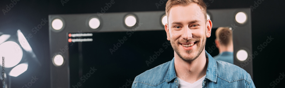 Fototapeta premium Panoramic shot of makeup artist smiling at camera near mirror in photo studio
