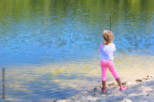 Fototapeta Naklejka Na Ścianę i Meble -  Little child fishing in summer on river on sandy shore against background of blue water and displaying green trees in river