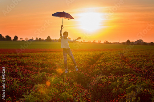 Fototapeta Naklejka Na Ścianę i Meble -  Beautiful child, flying with umbrella over gorgeous crimson clover field on sunset