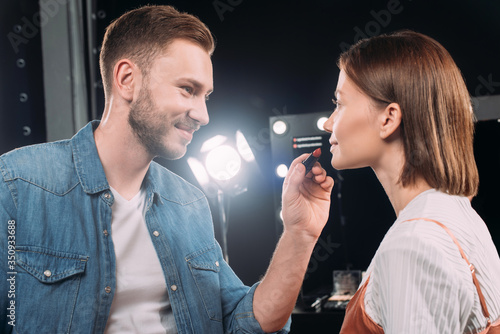 Handsome makeup artist smiling while applying lipstick on attractive model