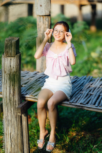Asian cute girl with glasses wear pink shirt and white pants sitting on wooden bridge in garden at home. © Pornthiwa