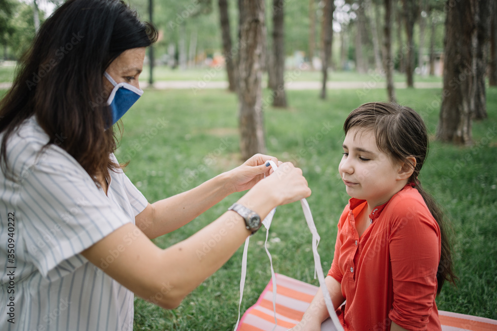 Mother with mask putting mask to her daughter outdoor. Woman wearing medical mask setting mask to her kid while sitting in park.