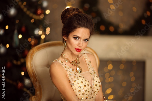 Beauty portrait of elegant woman with wedding hairstyle and makeup. Beautiful brunette girl with golden jewelry in prom dress sitting on modern chair over bokeh lights xmas decorations.