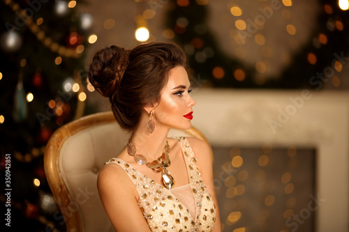 Luxurious portrait of elegant woman with wedding hairstyle and makeup. Beautiful brunette girl with golden jewelry in prom dress sitting on modern chair over bokeh lights xmas decorations.