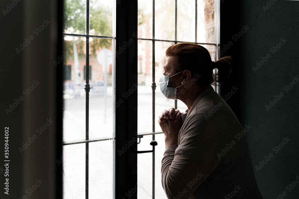 An older woman is looking through the window. She is wearing a protective mask on her face. She is protecting herself and staying at home. She has her hands joined on her chest. Pandemic concept. An older woman is looking through the window. She is wearing a protective mask on her face. She is protecting herself and staying at home. She has her hands joined on her chest. Pandemic concept.