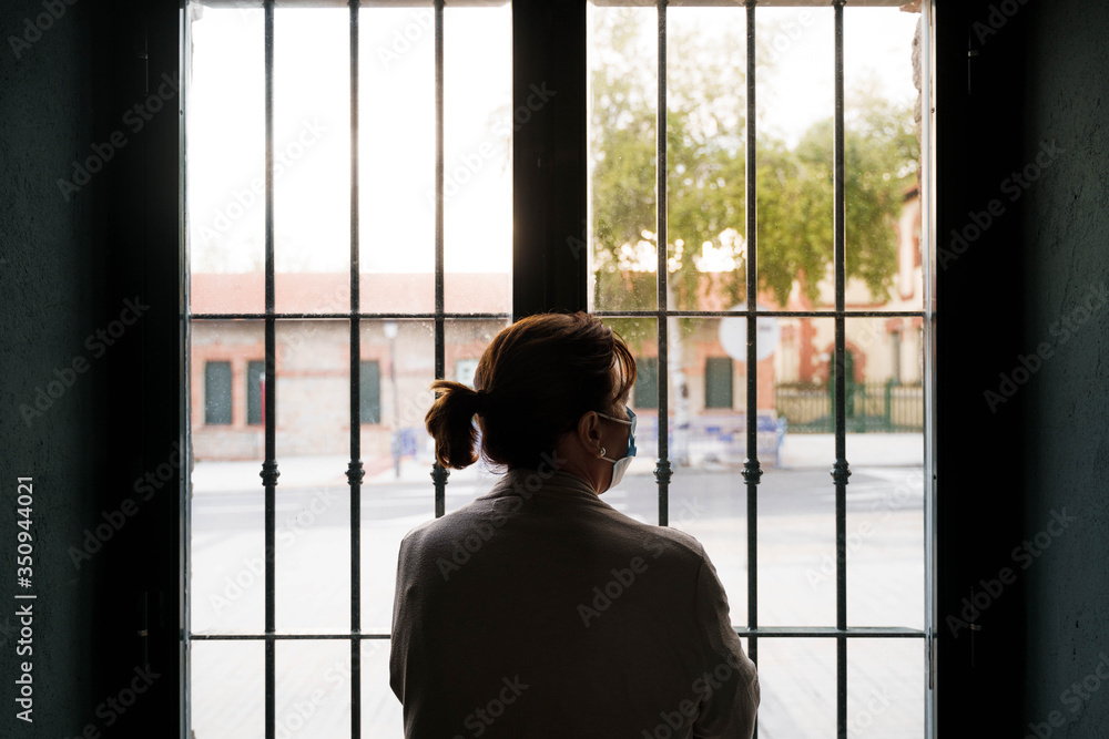 An older woman is looking through the window. She is wearing a protective mask on her face. She is protecting herself and staying at home. Pandemic concept. An older woman is looking through the window. She is wearing a protective mask on her face. She is protecting herself and staying at home. Pandemic concept.