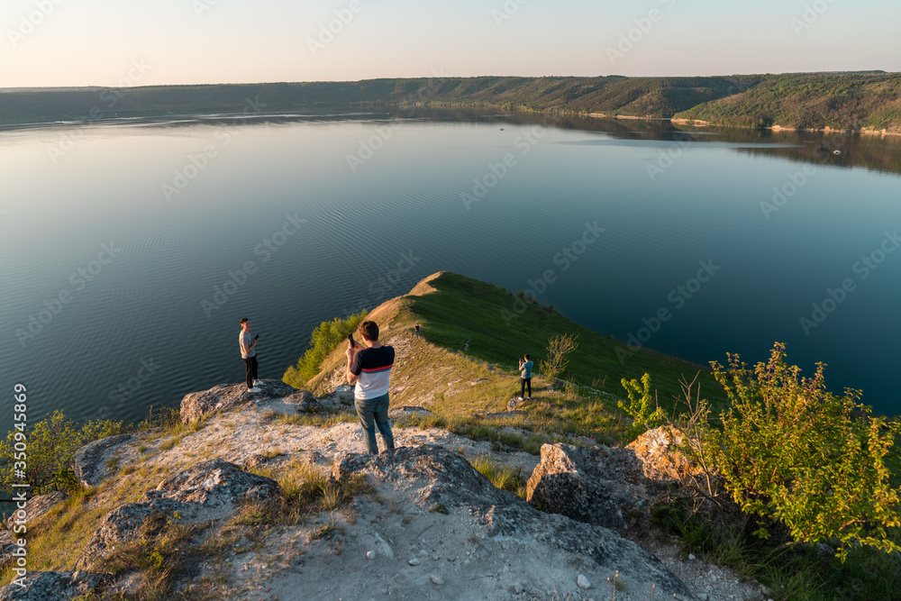 Young friends with phones standing on the top of rock and looking at the wide river sunset in summer