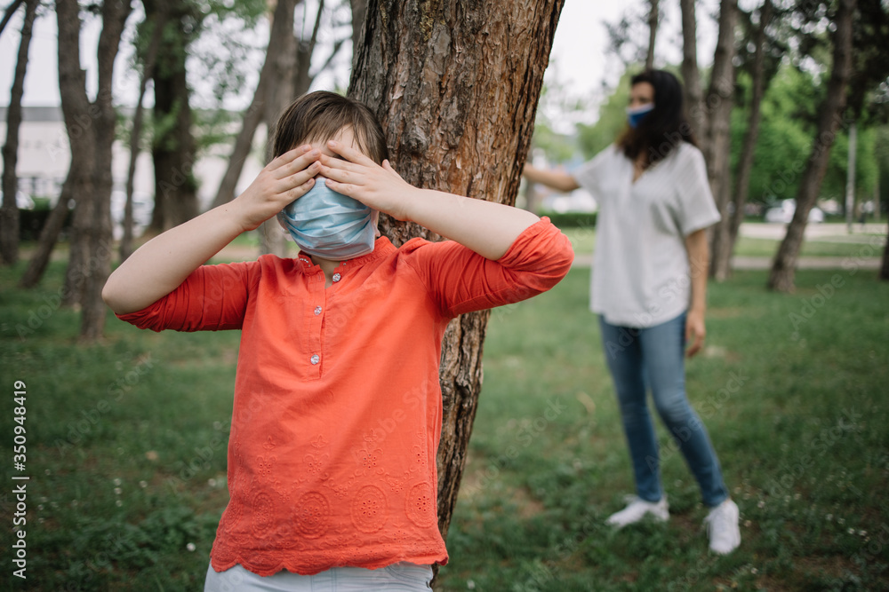 Girl wearing medical mask hiding her eyes with hands while counting. Mother and child playing hide-and-seek while wearing antivirus masks during coronavirus pandemic.