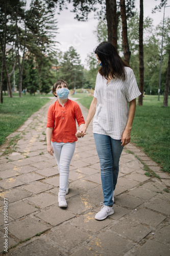Woman holding little girl's hand while walking in park. Mom and child walking on park alley while wearing antivirus masks during coronavirus pandemic.
