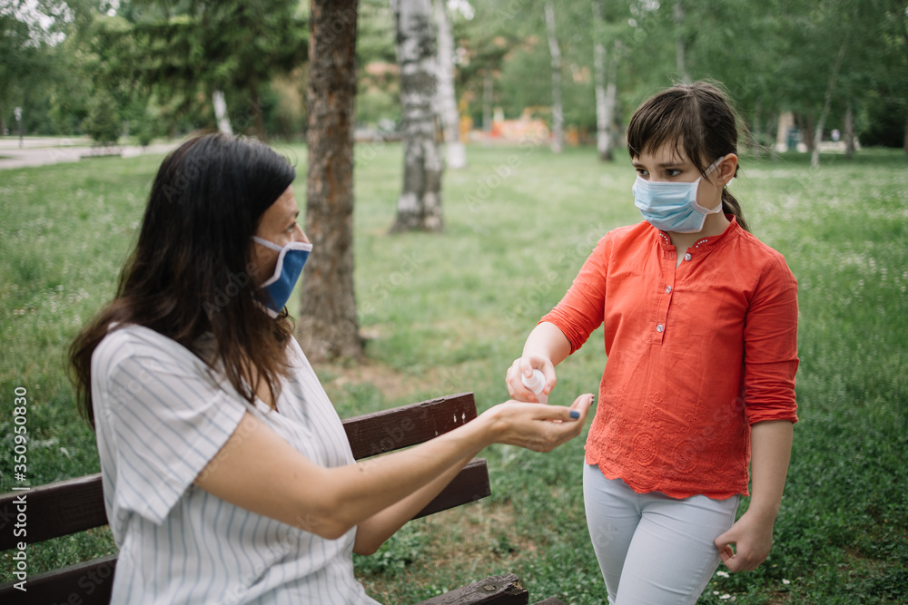 Fototapeta premium Little girl with mask holding disinfectant over woman's hands outdoor. Cute girl with antivirus mask holding hand sanitizer while standing in park near her mom who is sitting on bench.