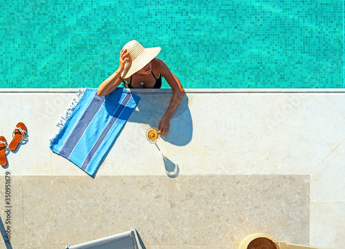 Top view from above of turqiuose swimming pool with young woman, bikini, hat and martini glass in blue water background with copy space