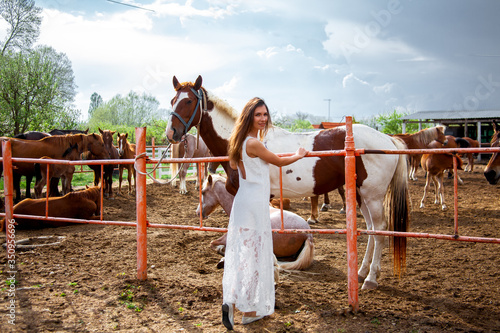 Beautiful woman dressed white dress, walking outdoor near horse paddock in the ranch. Friendship lifestyle of woman with wild animal in the nature