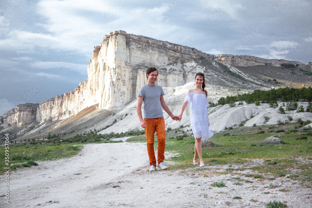 Happy couple in relationship walking in the beautiful place. Female dressed white dress and posing against the white mountain cloudy sky. Man hugging his girlfriend