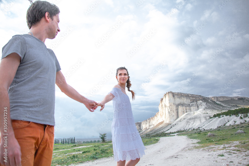 Happy couple in relationship walking in the beautiful place. Female dressed white dress and posing against the white mountain cloudy sky. Man hugging his girlfriend Happy couple in relationship walking in the beautiful place. Female dressed white dress and posing against the white mountain cloudy sky. Man hugging his girlfriend