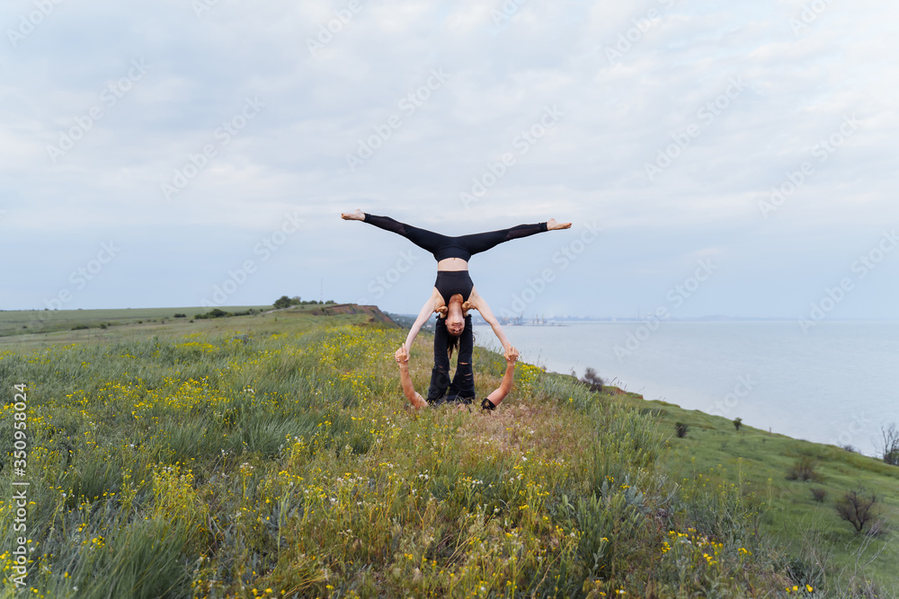 Attractive couple practicing yoga outdoors on the shore of the sea. Girl doing splits, being upside down during acroyoga, lying on the legs of a man. Sport exercises on the hill. Long distance shot Attractive couple practicing yoga outdoors on the shore of the sea. Girl doing splits, being upside down during acroyoga, lying on the legs of a man. Sport exercises on the hill. Long distance shot