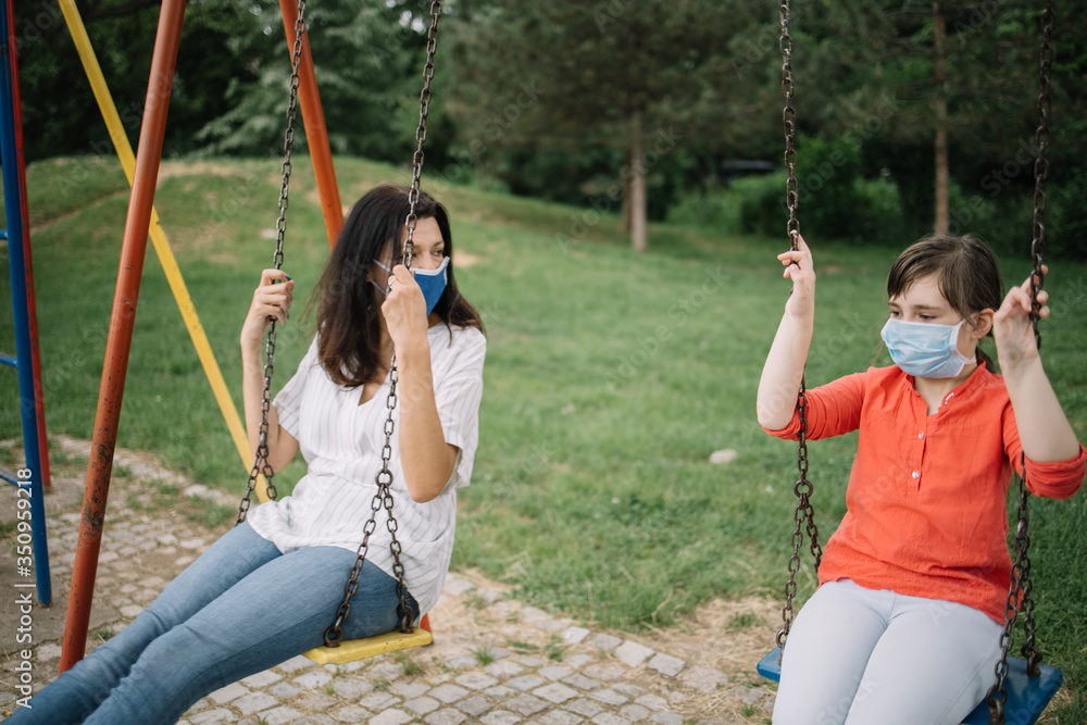 Pretty girl and woman wearing protection mask while enjoying playground facilities. Young girl and her mother swinging at the playground while wearing mask