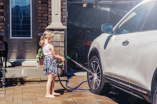 Young preschool girl washing car on driveway in front house on summer day. Kids home errands duty chores responsibilities concept. Child playing with a hose spraying water. Home work lifestyle.