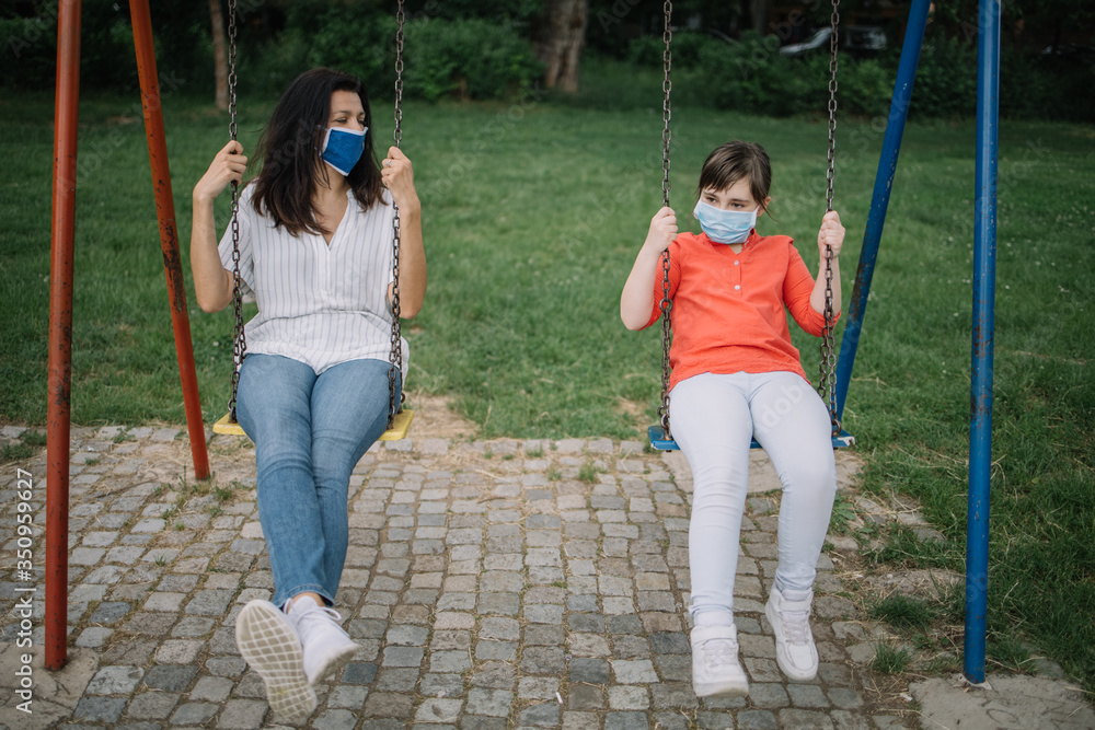 Portrait of girl with antivirus mask swinging in park with adult woman. Lady with protection mask swinging with her daughter during coronavirus pandemic.