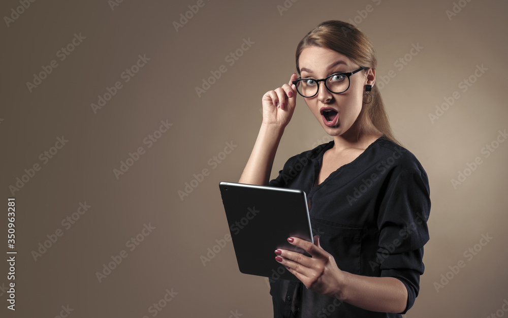 Girl wearing eye glasses and holding digital tablet with shocked face on beige background with copy space Girl wearing eye glasses and holding digital tablet with shocked face on beige background with copy space