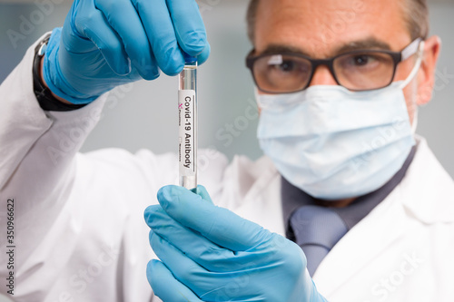 Scientist or doctor with medical face mask and medical gloves is handling virus test tube with antibody result markers in front of laboratory in background
