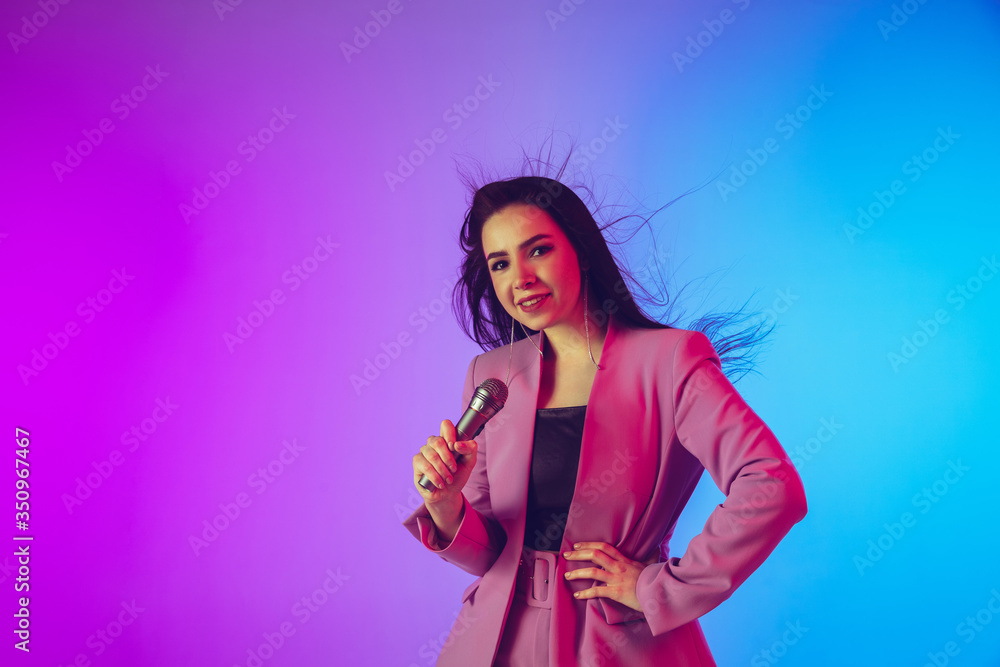 Naklejka premium Caucasian female singer portrait isolated on gradient studio background in neon light. Beautiful female model in pink wear with microphone. Concept of human emotions, facial expression, ad, music, art