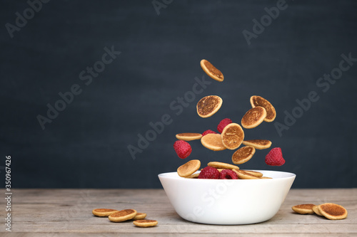 Flying tiny pancake cereal - trendy quarantine food. Mini pancakes and fresh raspberries in motion falling in white bowl on black chalkboard background. Levitation, gravity. Homemade breakfast.