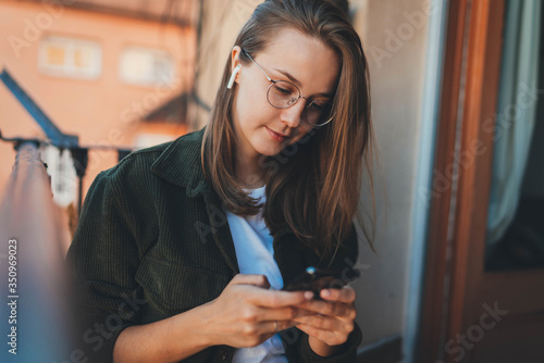 Charming young woman in trendy eyeglasses ordering food online via app on smartphone while working from home, Millennial hipster girl smiling texting message to friends in social networks