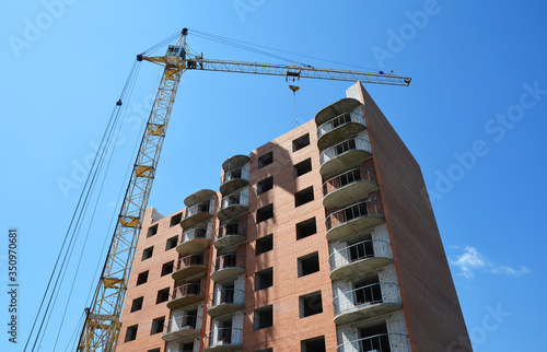 Obraz na plátně A new brick high-rise residential building is constructed with the help of a tower, self erecting, saddle jib construction crane against blue sky