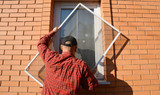 A handyman is installing anti-insect mosquito net, screen, fly and bugs protection on a plastic window of a brick house to keep the mosquitos away.