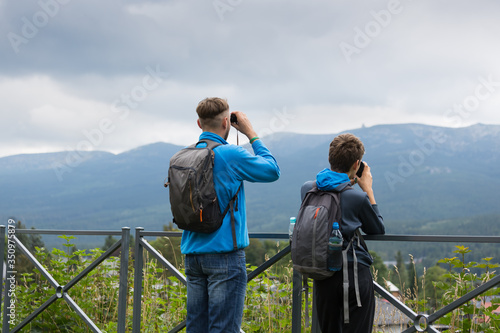 Mężczyzna i nastoletni chłopak  z plecakami oglądają przez lornetki odległy szczyt górski