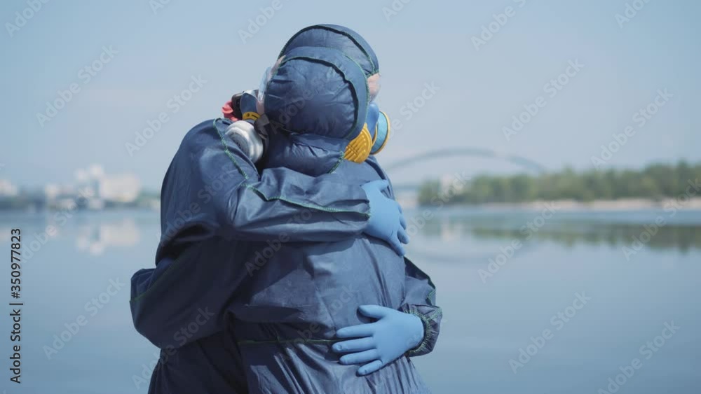 Unrecognizable woman in chemical suit coming to young loving man in respirator and protective eyeglasses outdoors. Young Caucasian couple hugging. Radioactive air pollution, Covid-19 pandemic. Unrecognizable woman in chemical suit coming to young loving man in respirator and protective eyeglasses outdoors. Young Caucasian couple hugging. Radioactive air pollution, Covid-19 pandemic.