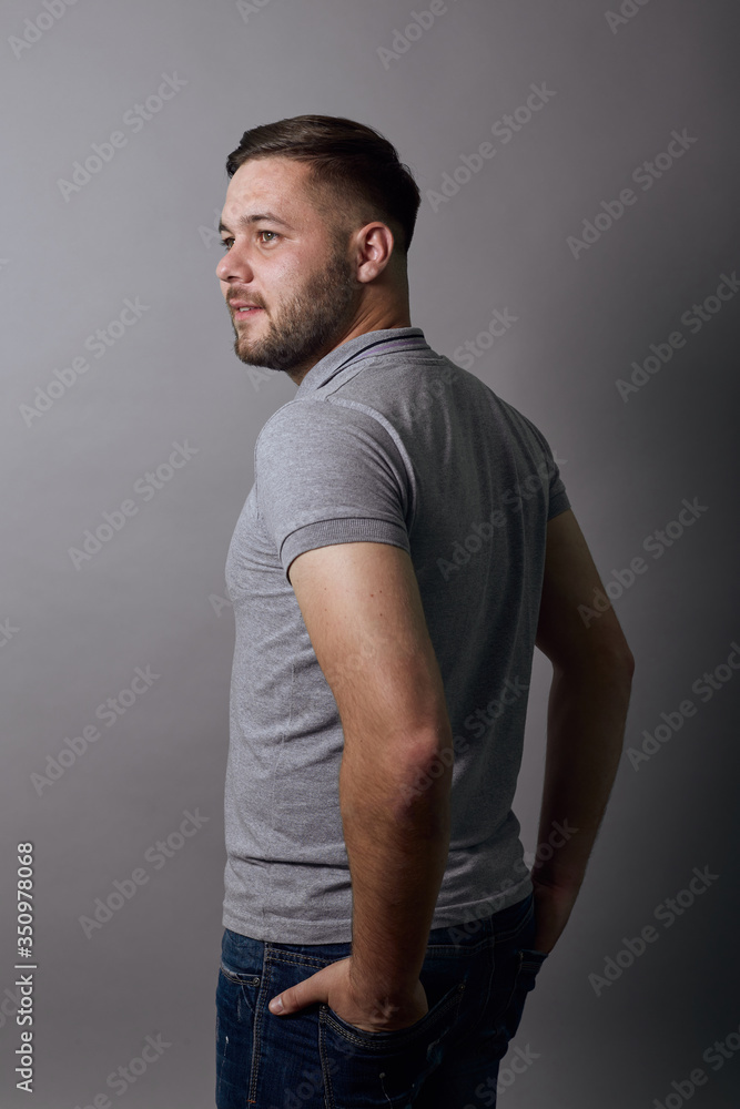 Young Bearded Dark Haired Man In Gray Stylish T-Shirt On Background, Portrait Of A Serious Male Who Looks Away. Young Bearded Dark Haired Man In Gray Stylish T-Shirt On Background, Portrait Of A Serious Male Who Looks Away.