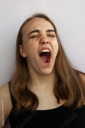 a young girl lazily yawns early in the morning, waking up and looking sleepy, tired and bored against a light wall, close-up
