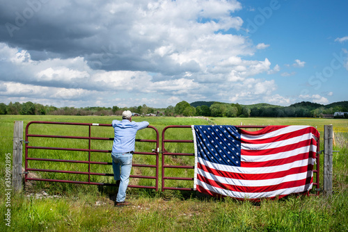 Farmer leaning on metal fence in grassy agricultural field, American flag draped over fence, blue sky white clouds