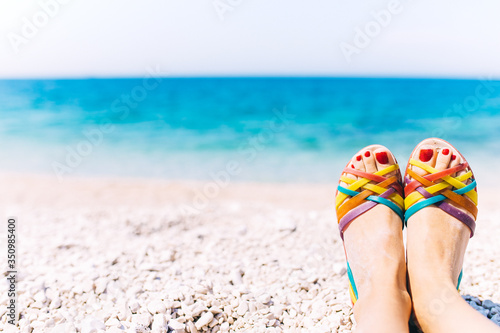 Selective focus of female legs and feet in beach rubber sandals on pebble beach with blurred turquoise sea background. Travel concept. Summer holidays. Vacation background with place for text