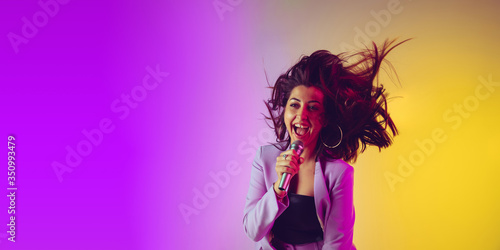 Caucasian female singer portrait isolated on gradient studio background in neon light. Beautiful female model in pink wear with microphone. Concept of human emotions, facial expression, ad, music, art