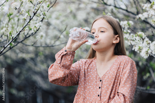 A little girl drinks clean fresh water from a plastic bottle near a flowering tree in the garden.