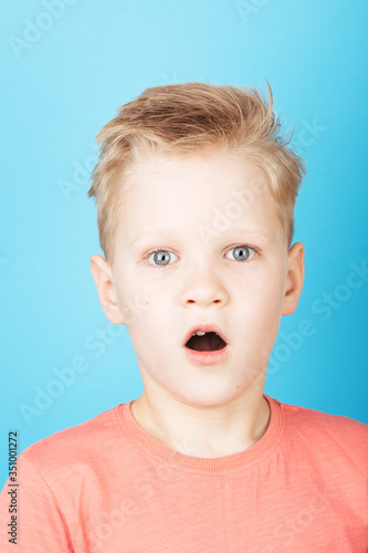 Portrait of cute surprised blondy boy with stylish hairstyle in coral T-shirt, closeup, on blue background.